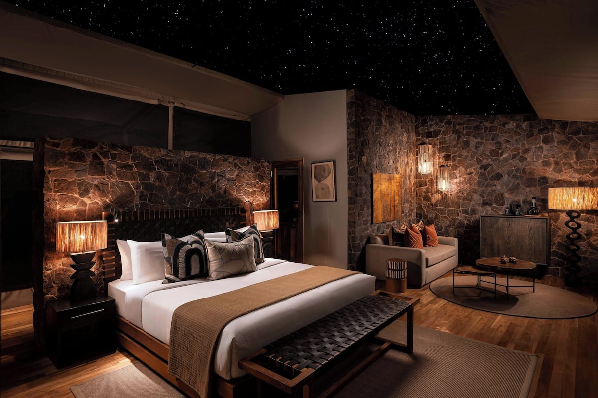 A bedroom with a stone wall, a bed with white bedding, a bench, and a nightstand with a lamp.
