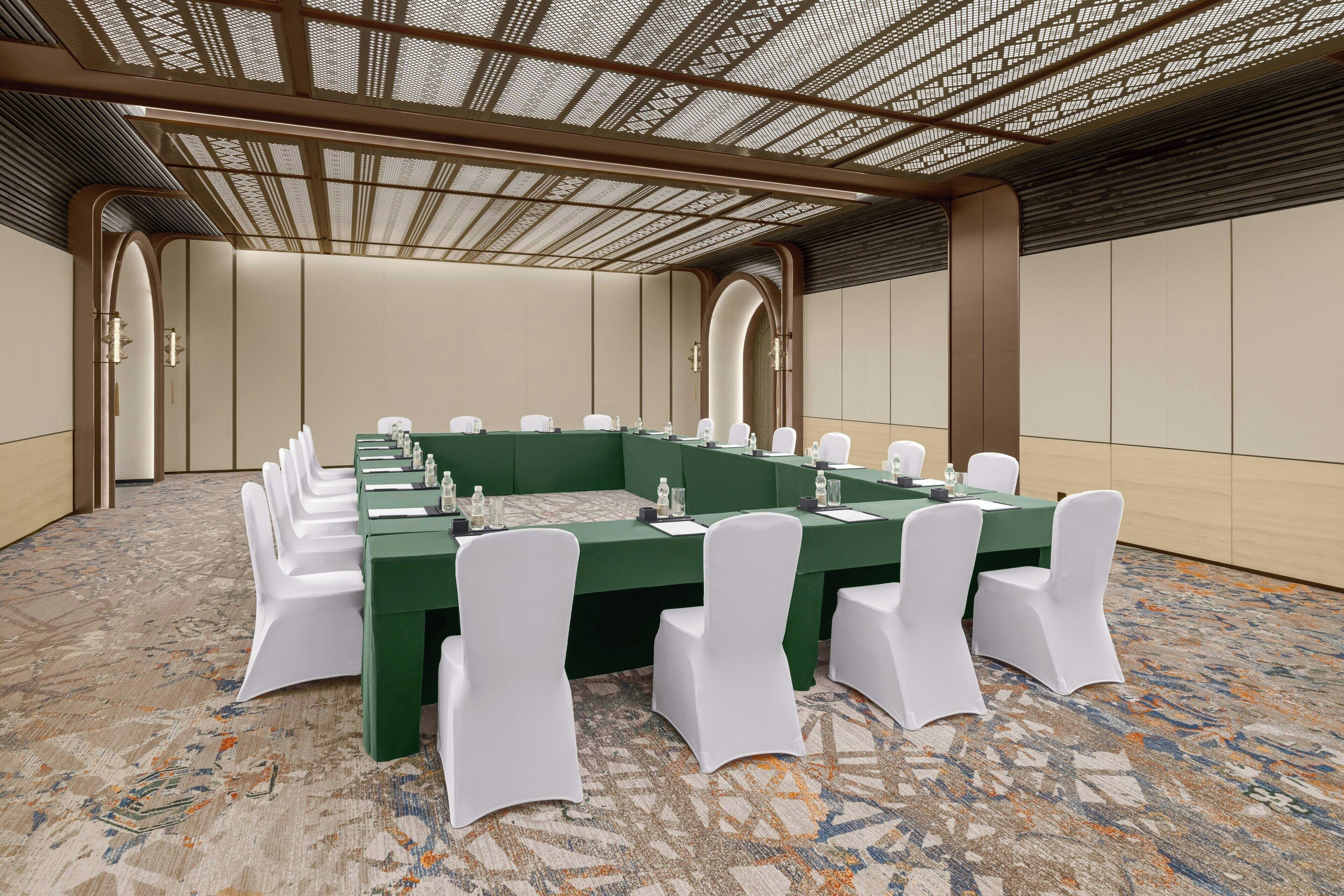 A conference room with a large oval table, white chairs, and a patterned carpet.