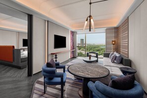 A modern hotel room with a sofa, chairs, a round table, a TV, and a large window offering a city view.