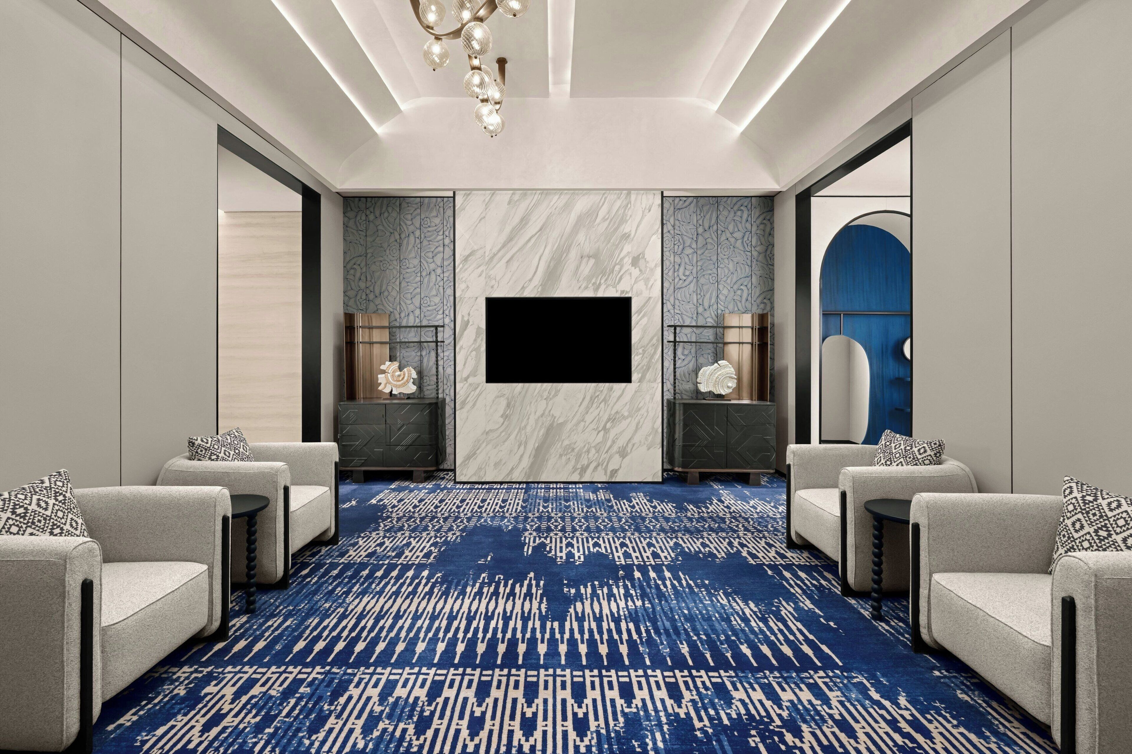 A modern hotel room with a large flat-screen TV, two armchairs, a patterned carpet, and a decorative wall panel.
