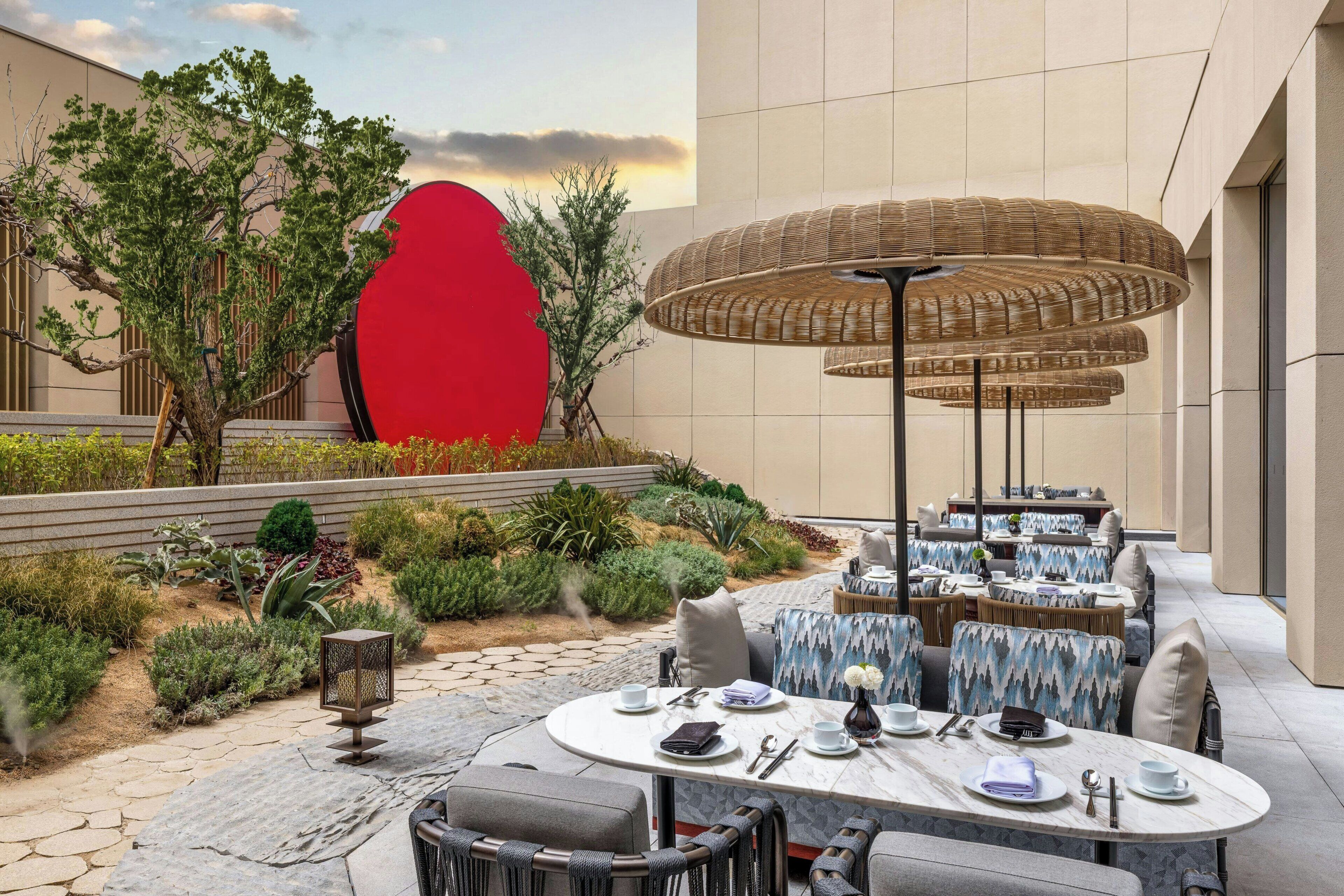 An outdoor dining area with round tables and chairs, surrounded by a landscaped garden with various plants and a large red sculpture.