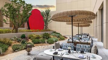 An outdoor dining area with round tables and chairs, surrounded by a landscaped garden with various plants and a large red sculpture.