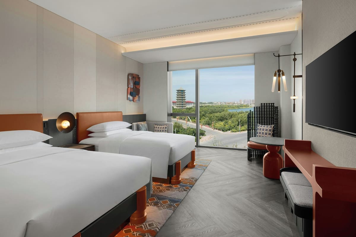 A hotel room with two beds, a desk, a chair, and a large window offering a view of a cityscape and greenery.