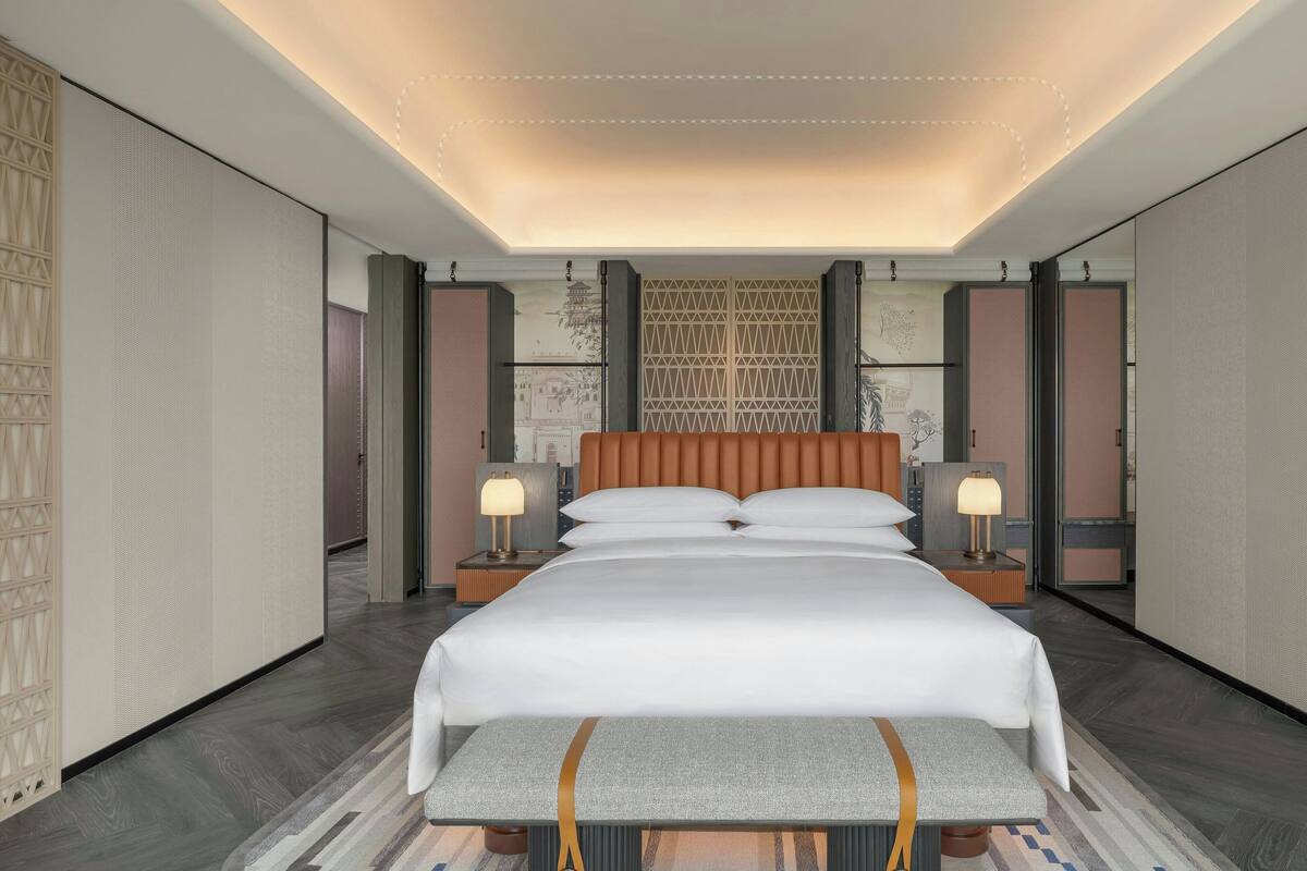 A modern hotel room with a large bed, two bedside tables with lamps, a bench at the foot of the bed, and a decorative wall panel.