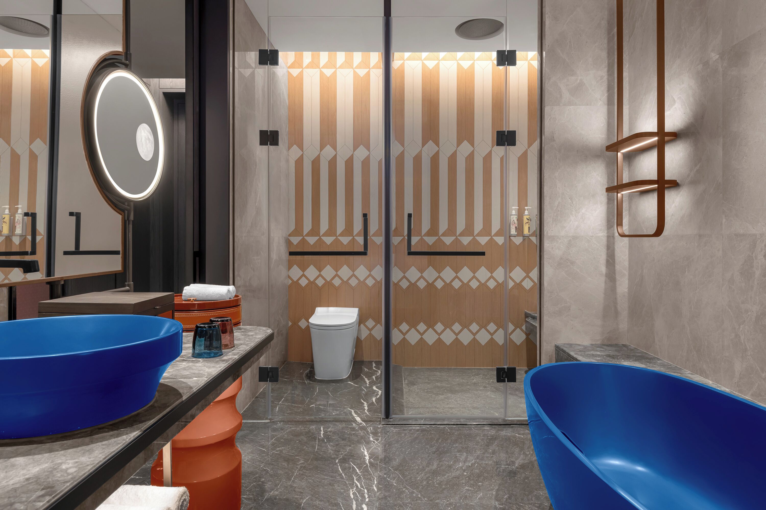 Un baño moderno con un lavamanos grande azul, un espejo redondo con iluminación y una zona de ducha con pared de diseño geométrico.