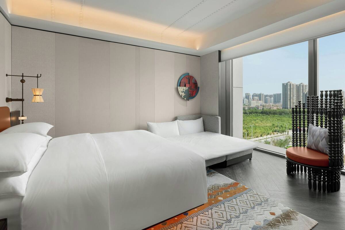 A modern hotel room with a large bed, a sofa, a view of the city, and a decorative wall piece.