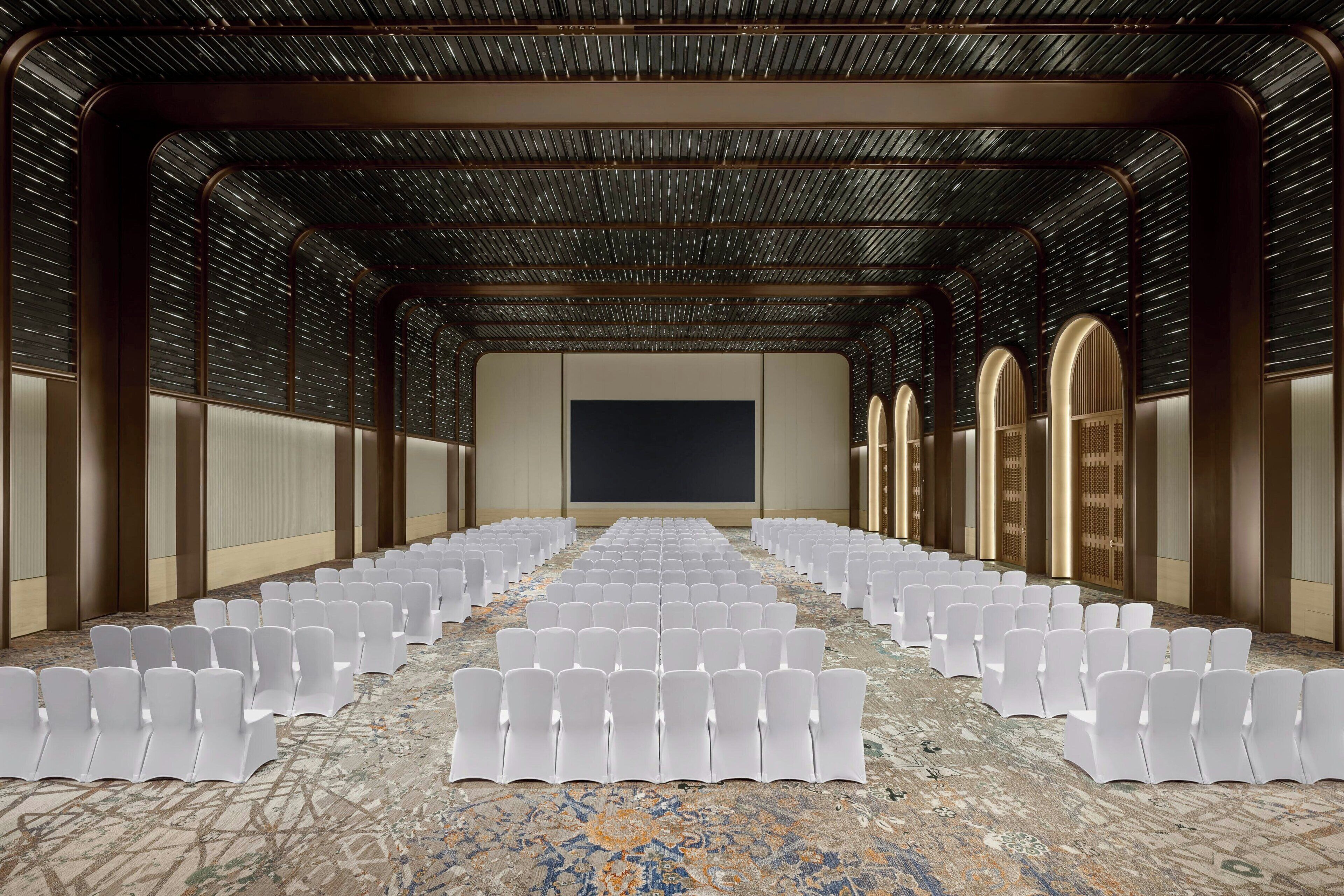 Un salón de conferencias con filas de sillas blancas, una pantalla grande y una alfombra estampada.