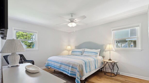 A bedroom with a bed, two bedside tables with lamps, a chair, a ceiling fan, and windows. - Rec Player's Retreat by Carolina Retreats (Carolina Beach)