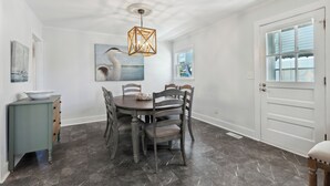 A dining area with a round table, six chairs, a decorative hanging light, and a door with a window. - Rec Player's Retreat by Carolina Retreats (Carolina Beach)