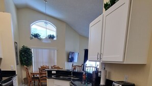 Una cocina con armarios blancos, encimera negra y suelo de baldosas. Hay un comedor con una mesa y sillas.