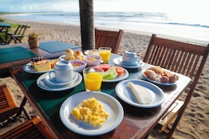 A breakfast table on a beach with plates of food, a glass of orange juice, and a view of the ocean. - Pousada Xalés de Maracaípe (Ipojuca)