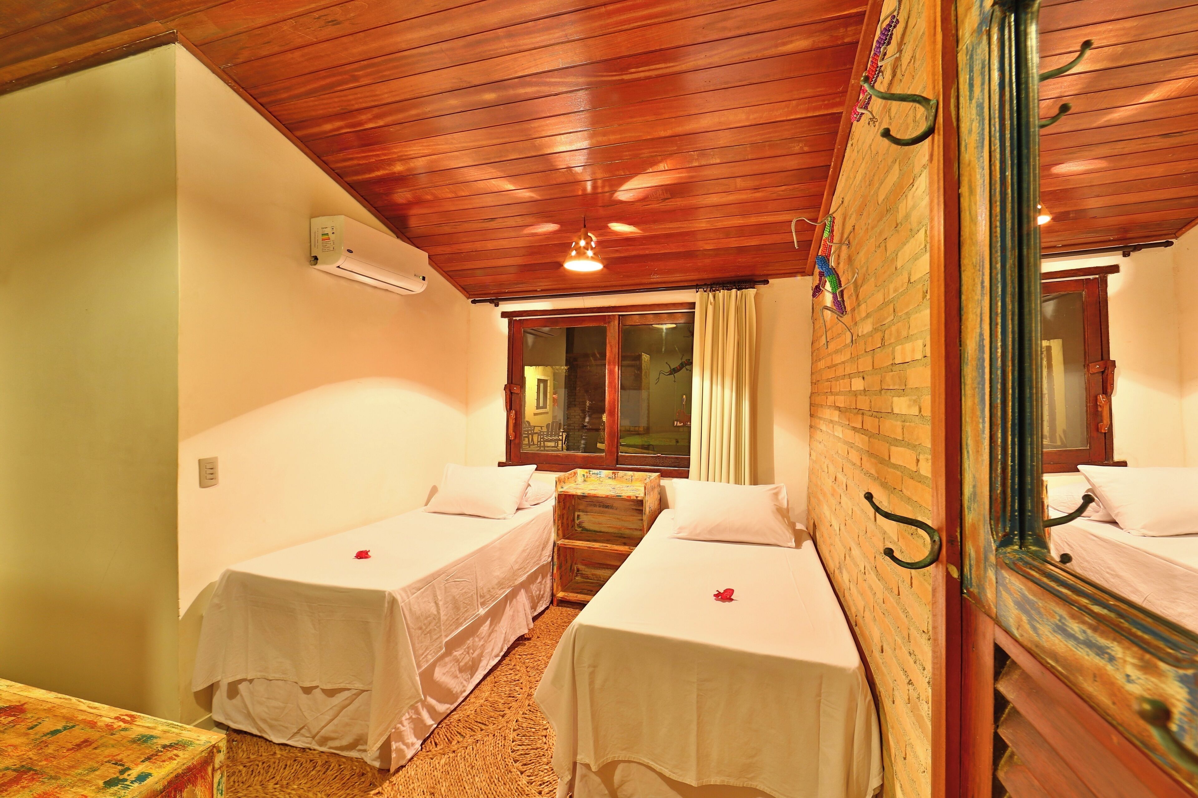 A room with two single beds, a wooden ceiling, a window with curtains, and a mirror on the wall.