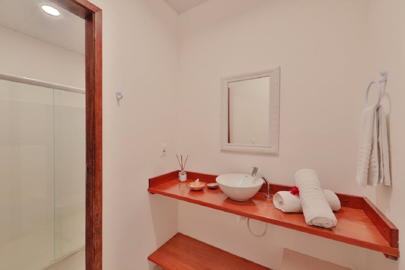 Un baño moderno con ducha de vidrio, lavamanos blanco, estante de madera con artículos de tocador y toallero.