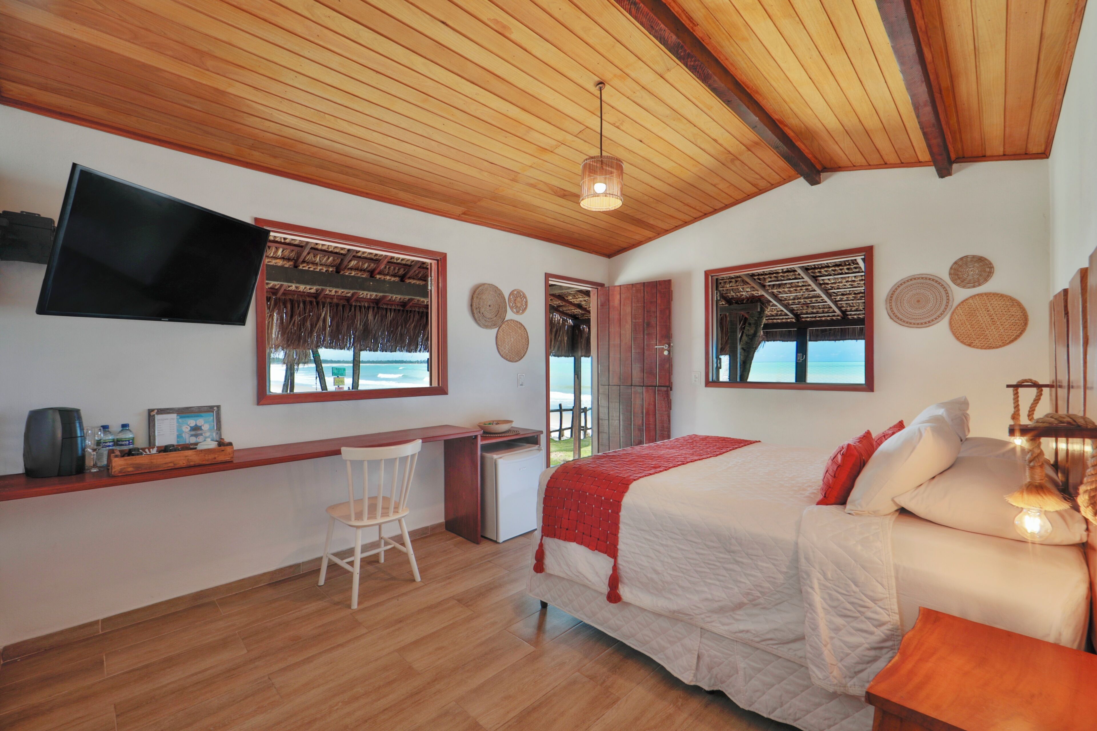 A bedroom with a wooden ceiling, a bed with a red blanket, a desk with a chair, and a TV mounted on the wall.