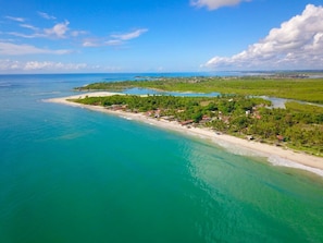 An aerial view of a coastal area with a beach, clear turquoise waters, and a small settlement with houses and greenery. - Pousada Xalés de Maracaípe (Ipojuca)