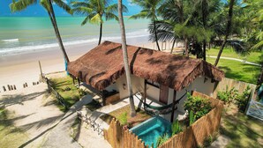 A thatched-roof structure with a pool, surrounded by a wooden fence and palm trees, overlooking a beach. - Pousada Xalés de Maracaípe (Ipojuca)