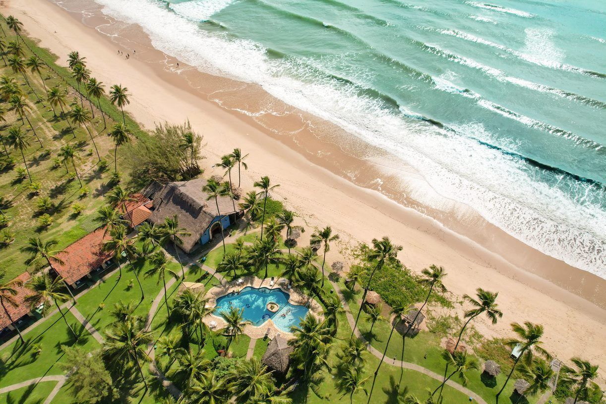 Vista aérea de un complejo turístico en la playa con piscina, palmeras y olas del océano.