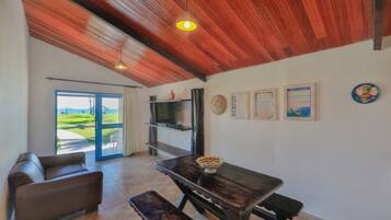 A spacious room with a wooden ceiling, a dining table with benches, a sofa, a TV on a shelf, and a view of the outdoors through a glass door.