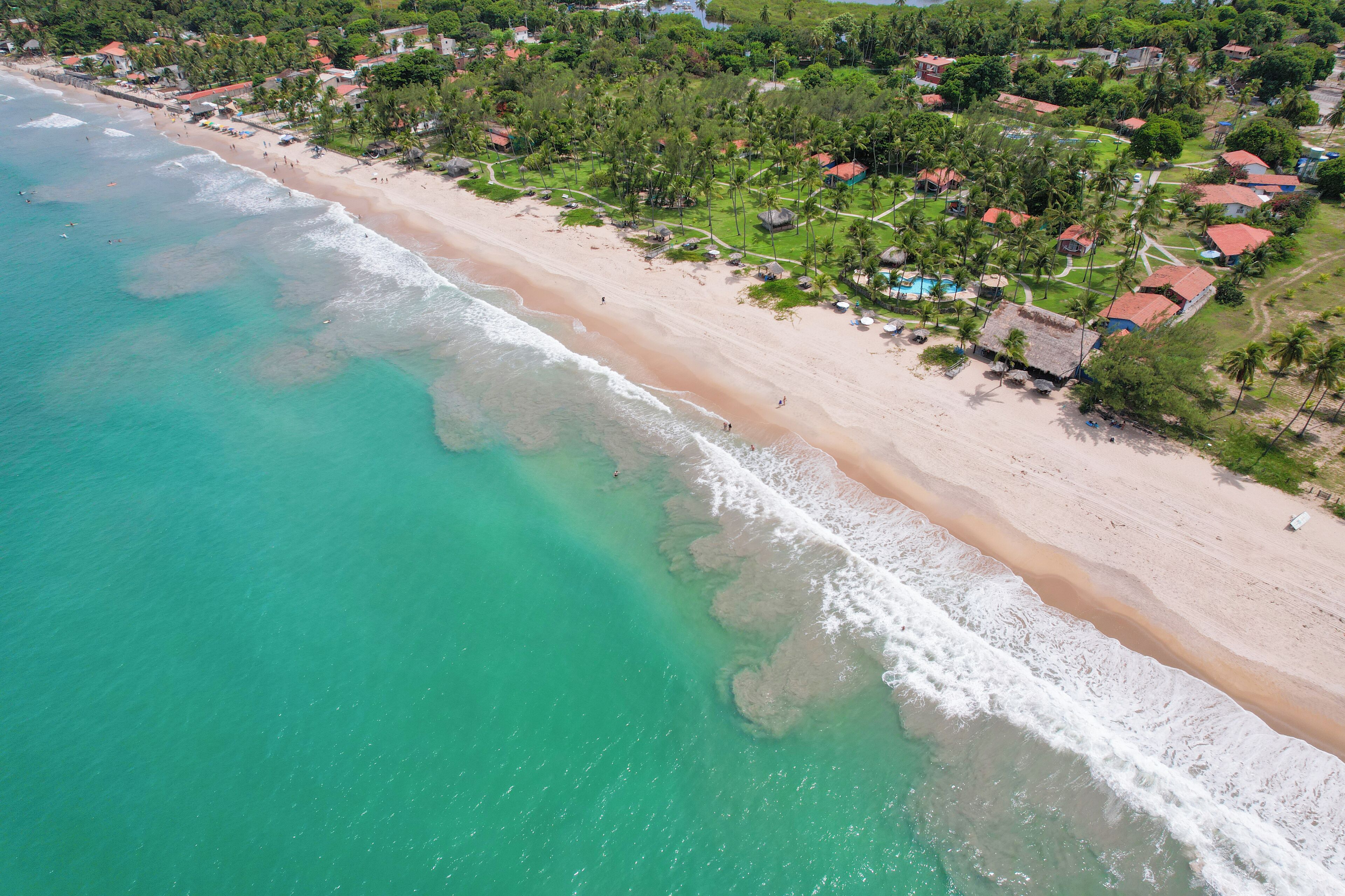 Vista aérea de un complejo turístico en la playa, con aguas turquesas cristalinas, una playa de arena y un conjunto de cabañas con techo de paja rodeadas de vegetación.