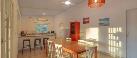 A spacious dining area with a wooden table and white chairs, a kitchen with a window, and a ceiling fan.