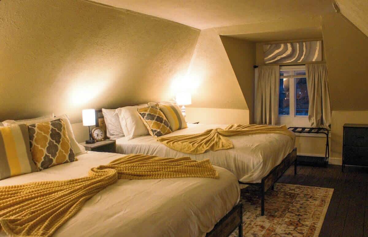 Ein Hotelzimmer mit zwei Betten, einem Nachttisch mit Lampe, einem Fenster mit Vorhängen und einem Kamin.
