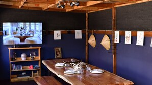 Ein Holztisch ist mit Tellern, Gläsern und Besteck gedeckt, ein Fernseher zeigt ein Zimmer und an der Wand hängen gerahmte Kunstwerke.