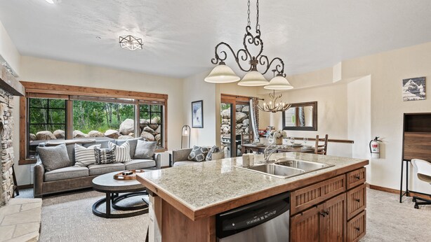 A spacious kitchen with a large island, wooden cabinets, and a dishwasher. There is a dining area with a round table and chairs, and a living room with a sofa and a fireplace.