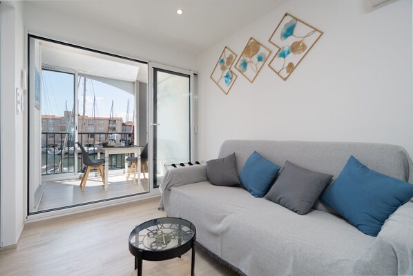 Una sala de estar moderna con un sofá gris, cojines azules, una mesa de centro de vidrio y vistas a un balcón con zona de comedor.