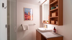 A bathroom with a glass shower door, a white sink, a wooden vanity, a towel rack, a framed picture, and a mirror with shelves.