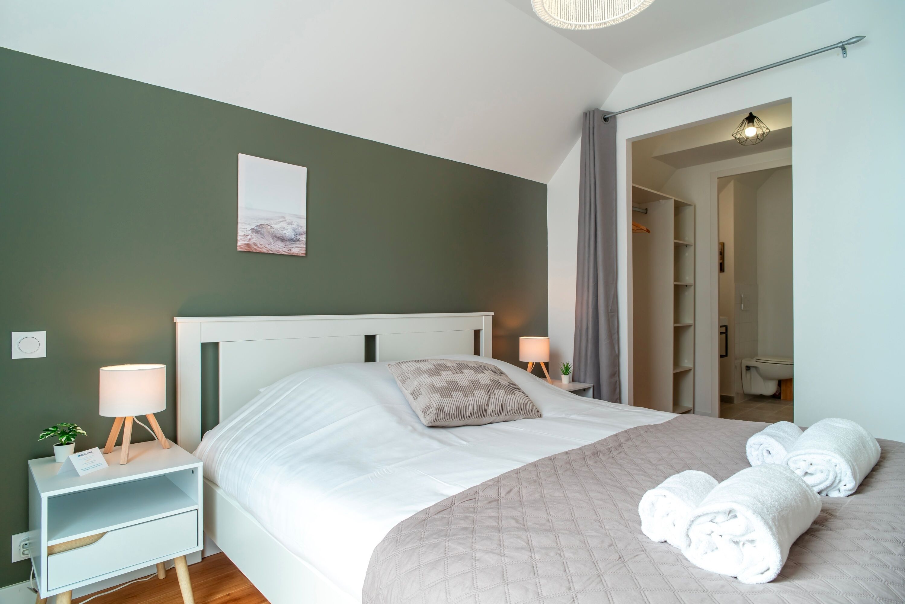 Un lit bien fait, recouvert d’une couverture matelassée et agrémenté de serviettes pliées, une table de chevet avec une lampe, une petite plante et un tableau encadré au mur.