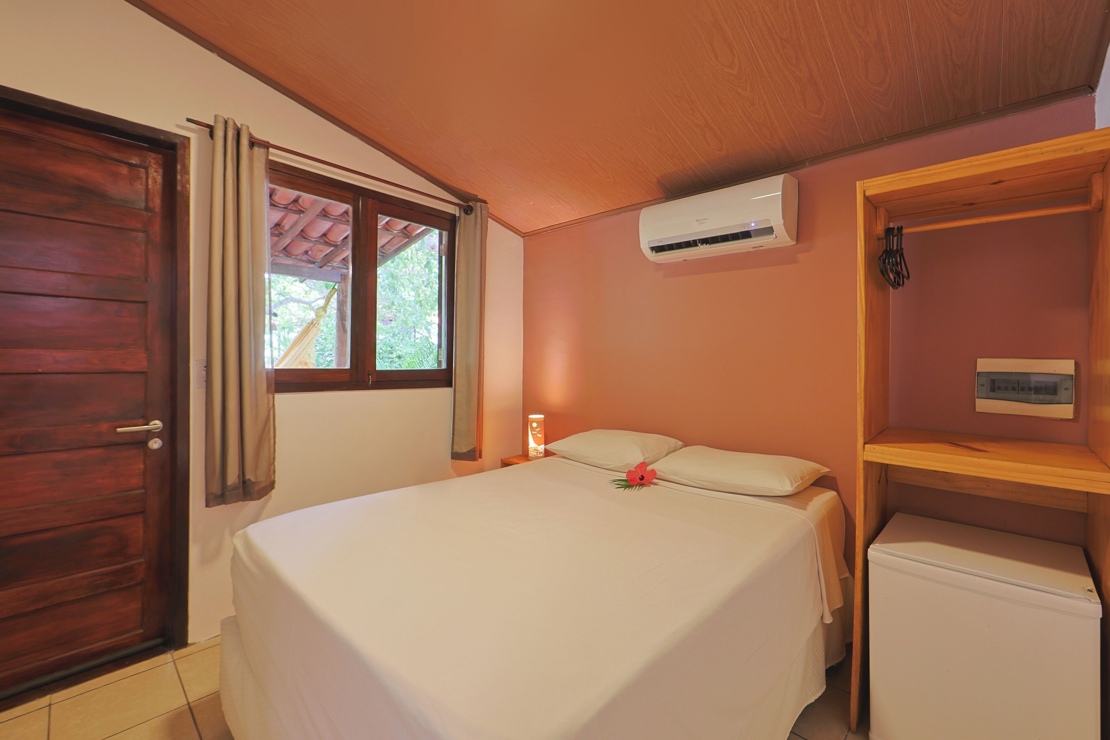 A bedroom with a bed, a wooden door, a window with curtains, an air conditioning unit, a bedside table with a lamp, and a small refrigerator.