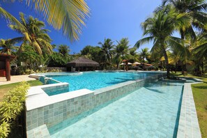 Una piscina con bordes de azulejos, rodeada de palmeras y una estructura con techo de paja.