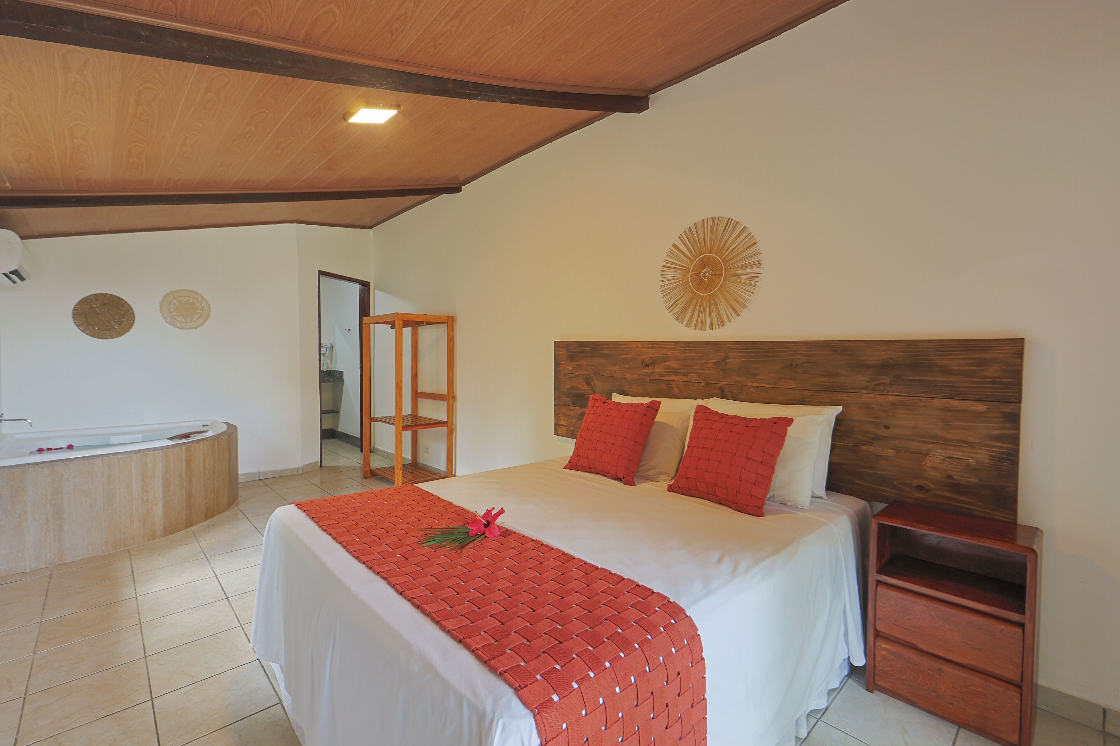 Een slaapkamer met een houten plafond, een bed met rode en witte beddengoed, een houten nachtkastje en een badkamer met een bad.