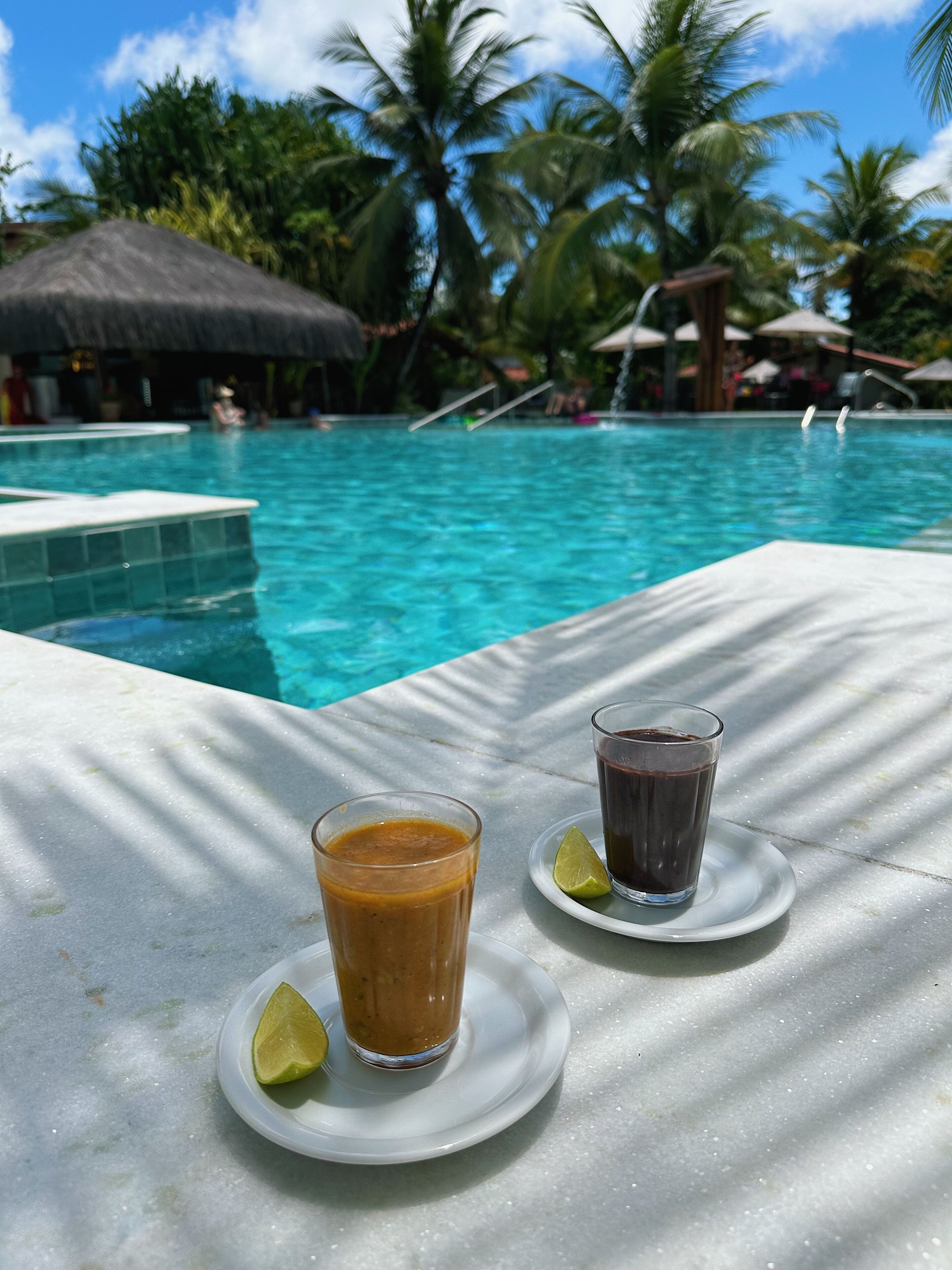 A poolside table with two drinks and a lime wedge, overlooking a swimming pool with palm trees and a thatched-roof structure in the background.