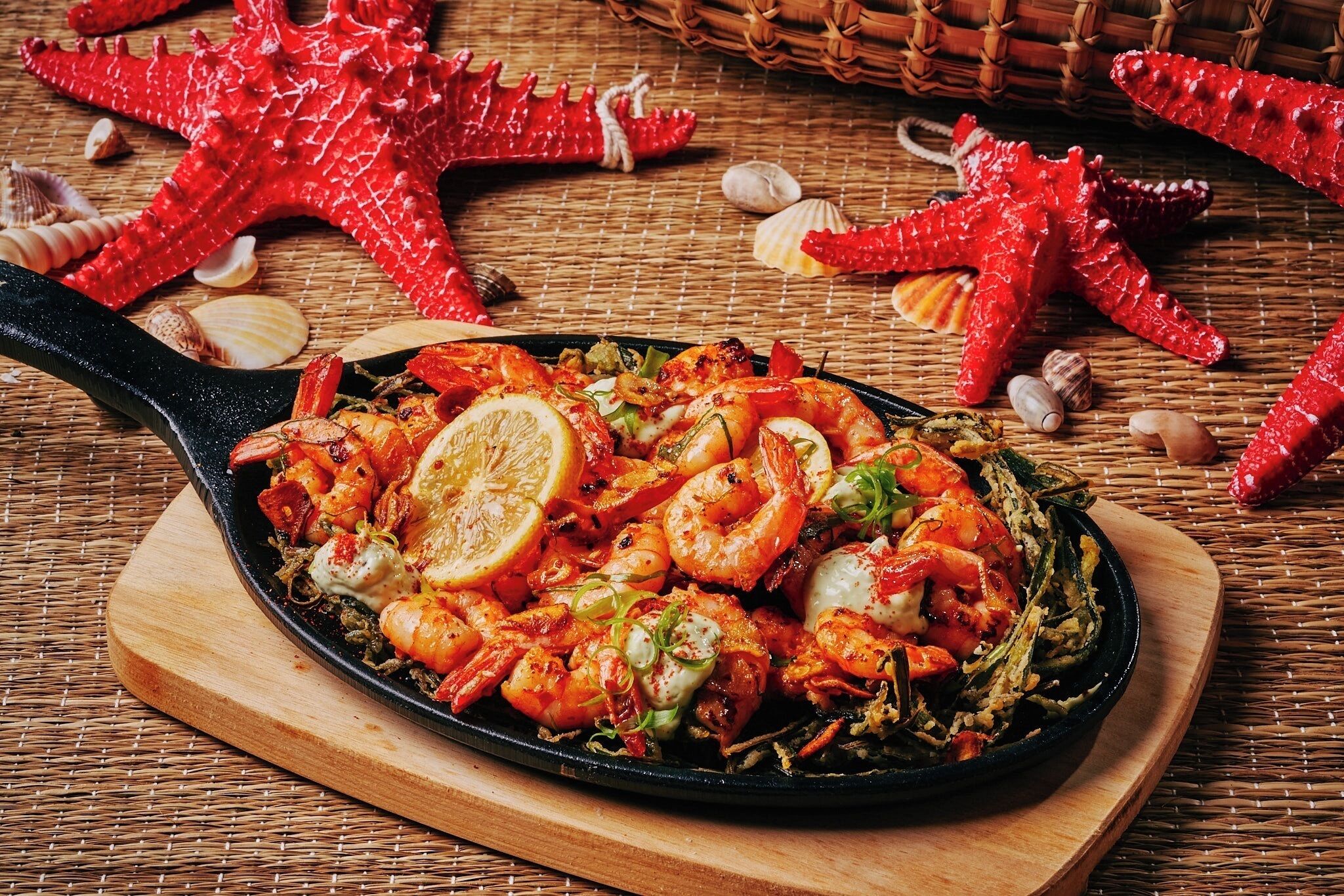 Una sartén con camarones y vegetales, rodeada de conchas marinas y estrellas de mar sobre una tabla de madera.