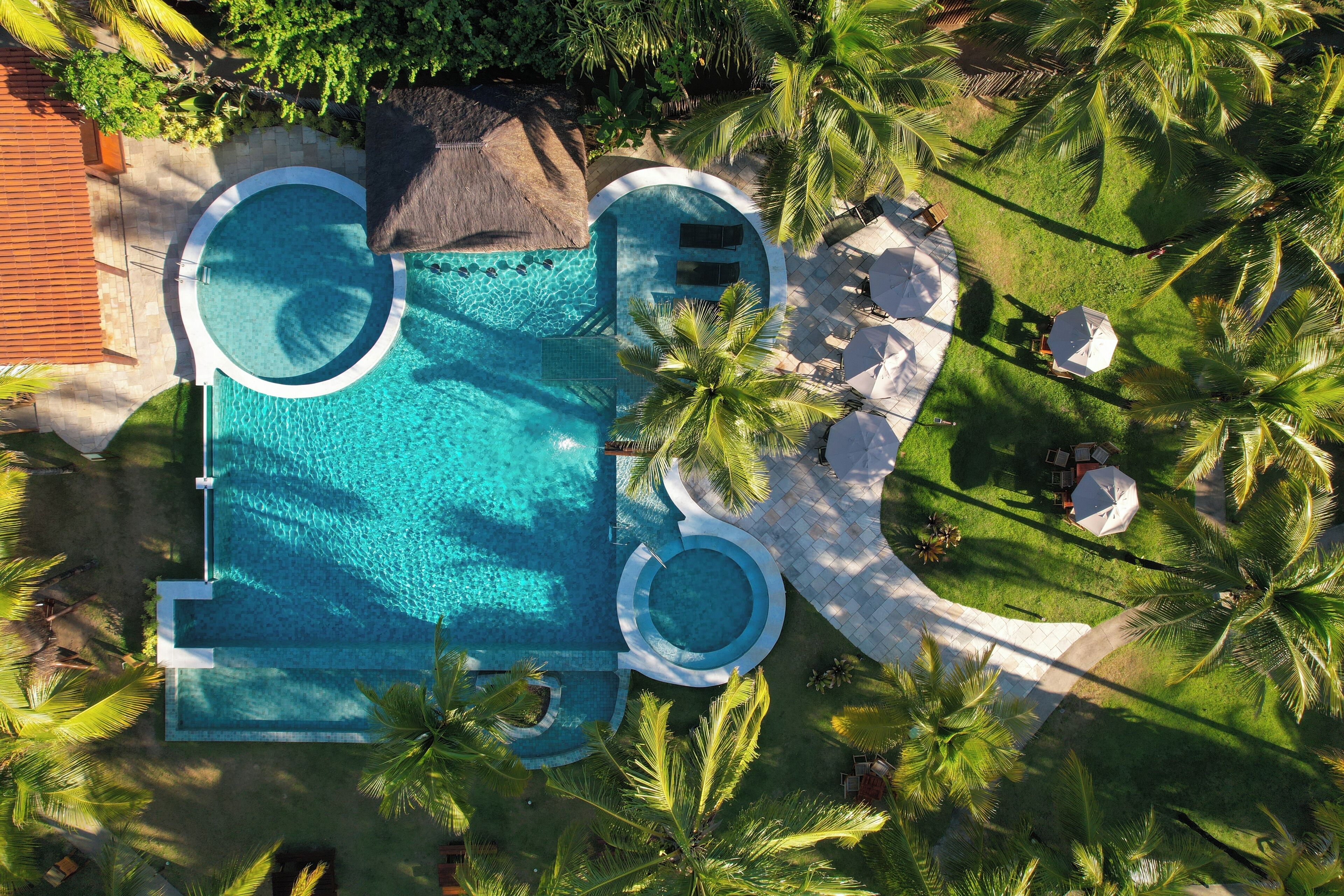 An aerial view of a resort with a large circular swimming pool surrounded by palm trees and lounge chairs.