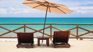 Deux fauteuils de plage en bois, avec un parasol beige, sur une plage de sable, donnant sur l’océan.