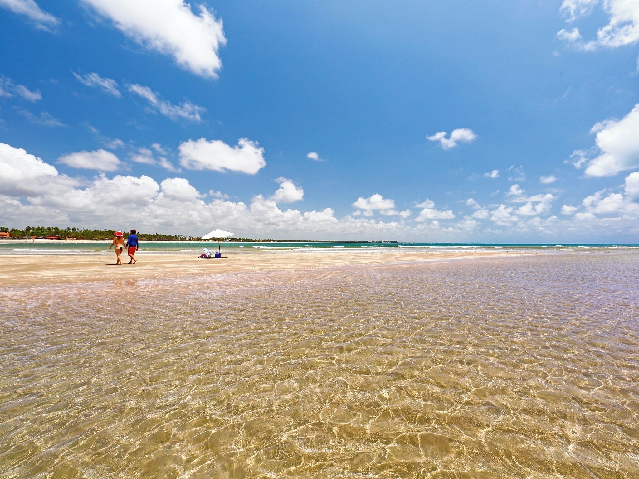 Ein Strand mit klarem, flachem Wasser, einigen Personen und einer entfernten Küstenlinie mit Gebäuden.