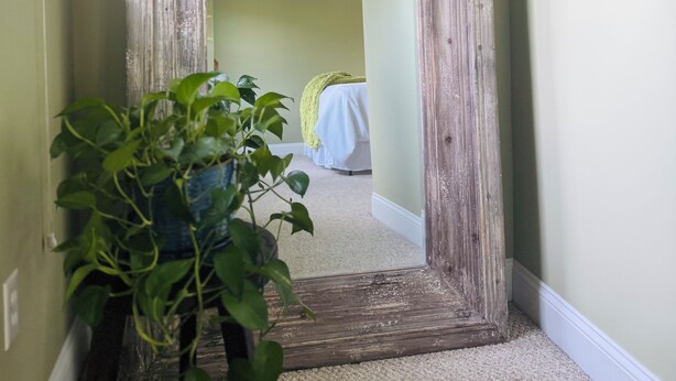 A hallway with a large, rustic mirror leaning against the wall, a potted plant in the foreground, and a glimpse of a bed with a yellow blanket in the background. - Lush Coastal Downtown Home - Beaches & Shopping (Freeport)
