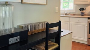 A kitchen with white cabinets, a dining table with a black chair, a framed picture on the wall, and a lamp on the table. - Lush Coastal Downtown Home - Beaches & Shopping (Freeport)