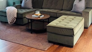 A living room with a sectional sofa, a coffee table, and a rug. There are two framed pictures on the wall and a lamp on a side table. - Lush Coastal Downtown Home - Beaches & Shopping (Freeport)