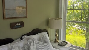 A bedroom with a bed, pillows, a bedside table with a lamp, a framed picture, and a window with a view of greenery. - Lush Coastal Downtown Home - Beaches & Shopping (Freeport)