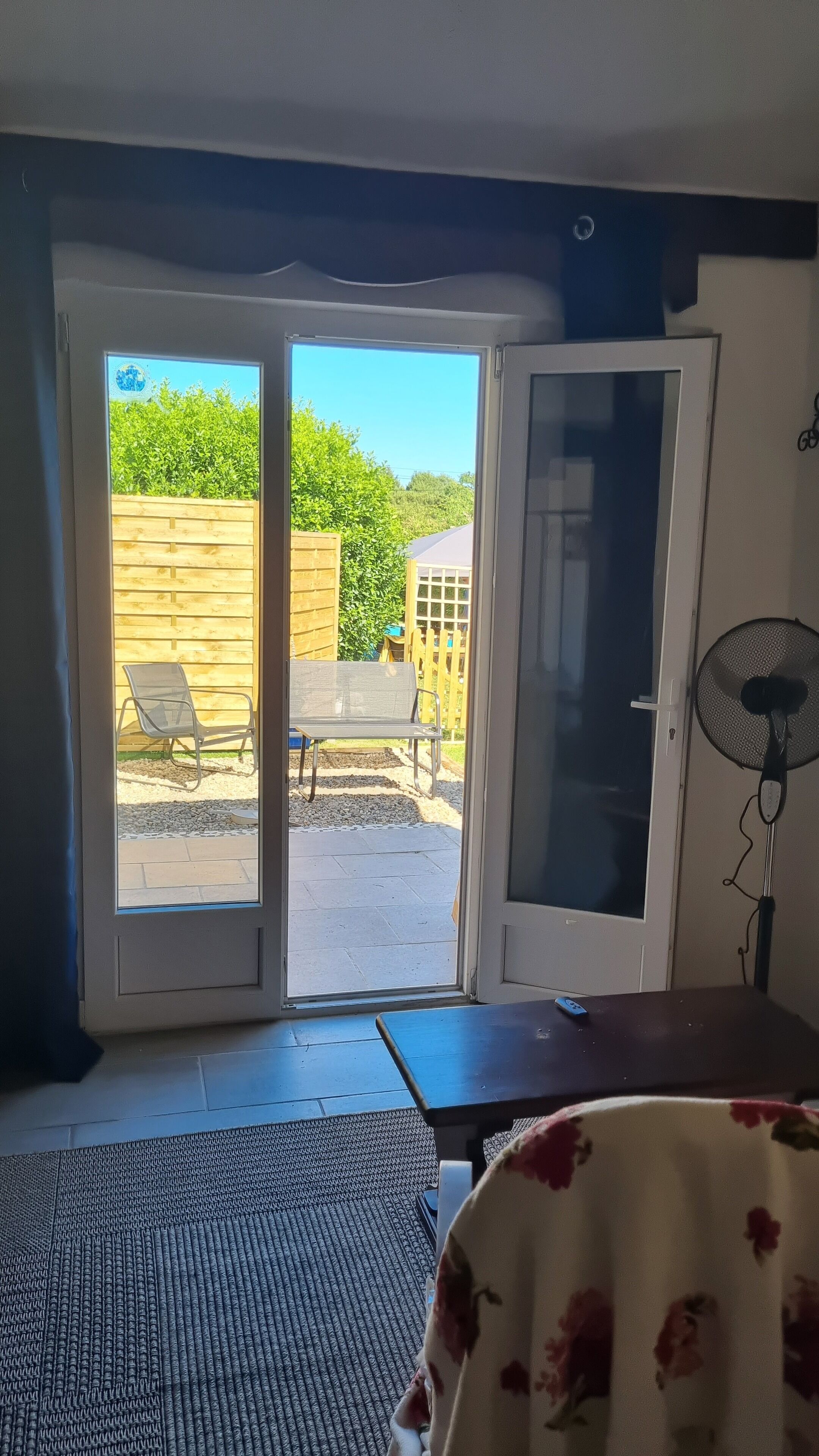 Ein Zimmer mit einer Schiebetür zu einer Terrasse, die von einem Holenzaun begrenzt wird, mit einer Bank und einem Ventilator.