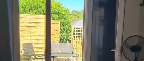 Ein Zimmer mit einer Schiebetür zu einer Terrasse, die von einem Holenzaun begrenzt wird, mit einer Bank und einem Ventilator.