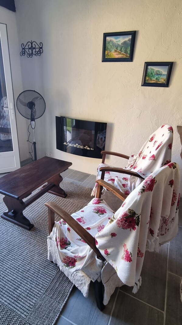 A cozy living room with a wooden chair covered in a floral blanket, a wooden table, a standing fan, and a fireplace. - Enchanting comfortable and private. (Bergerac)