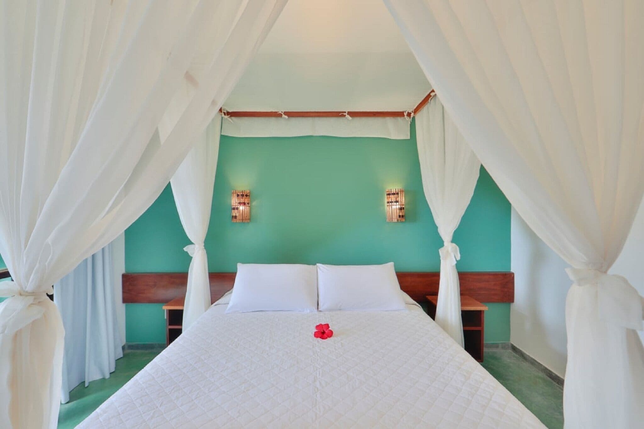 A bed with white bedding and a red flower, a wooden headboard, and a green wall with two wall sconces.