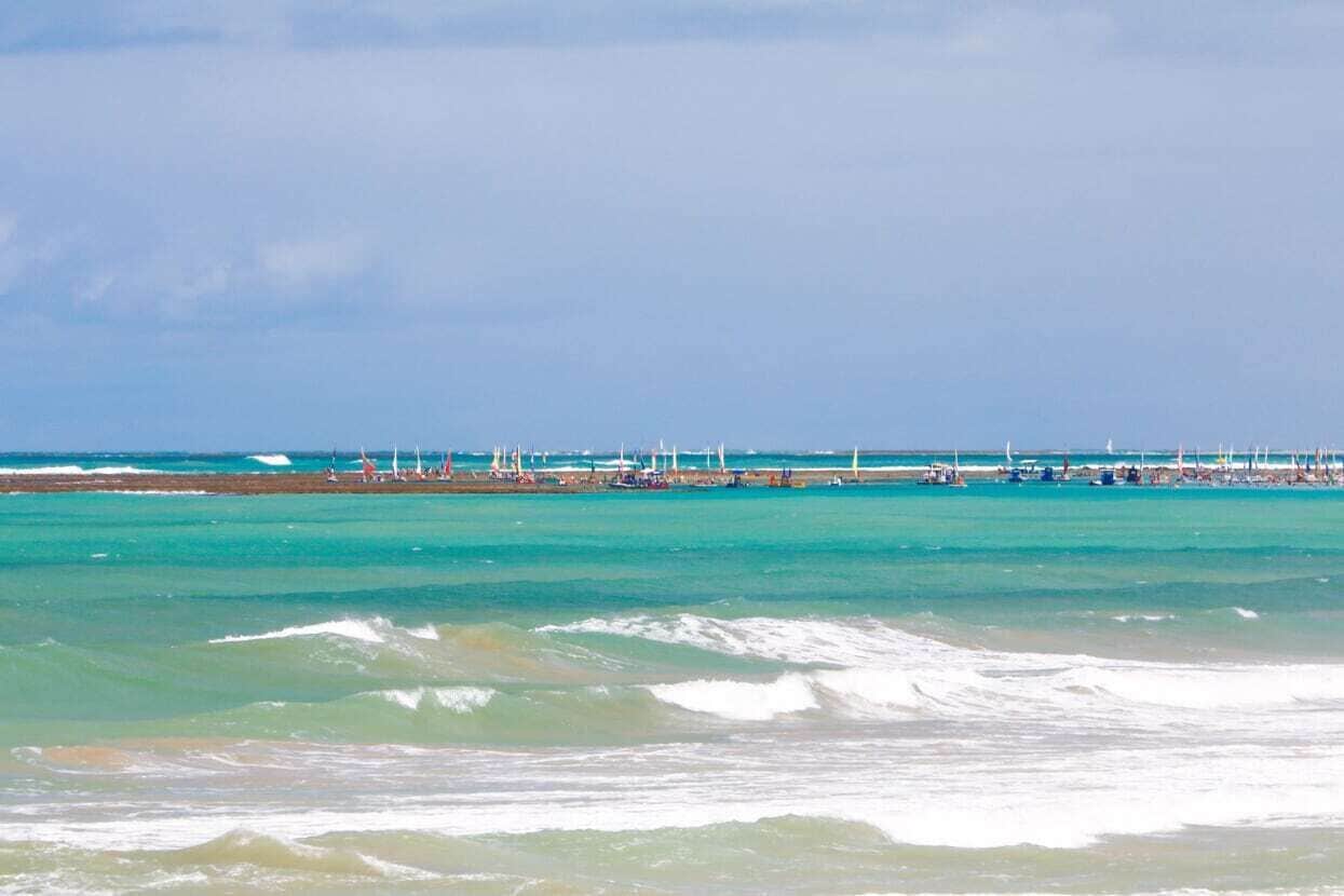 A coastal beach scene with clear turquoise waters, a sandy shore, and a row of sailboats anchored in the distance.