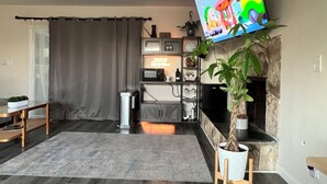 A modern living room with a flat-screen TV mounted on the wall, a fireplace, a kitchen area with a microwave and shelves, and a large potted plant.