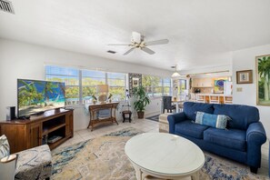 A spacious living room with a large TV, a blue sofa, a white coffee table, and a ceiling fan.