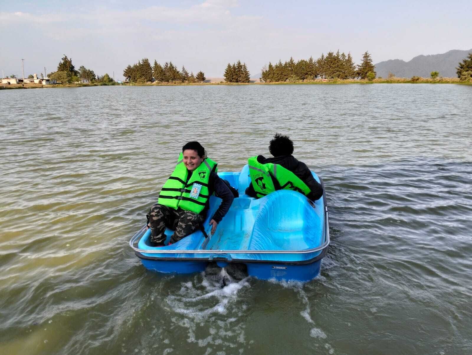 Two people in a blue inflatable boat on a lake with trees and buildings in the background.
